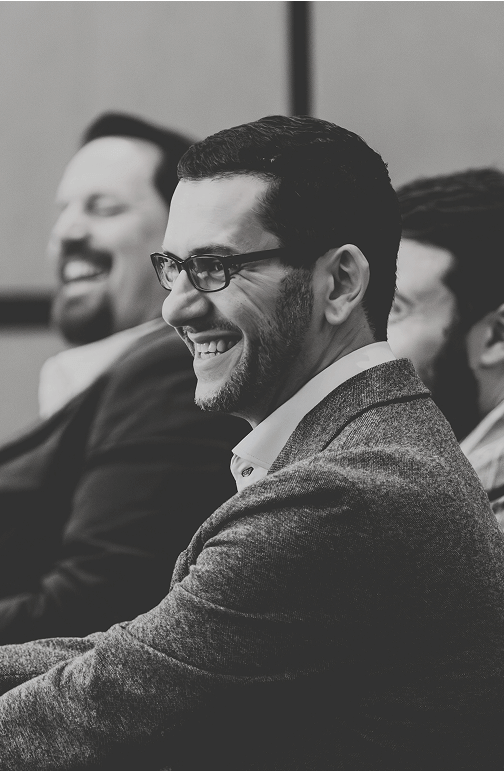 Black and white photo of a smiling man in glasses and business attire, engaged in a conference session with colleagues laughing in the background Black and white photo of a smiling man in glasses and business attire, engaged in a conference session with colleagues laughing in the background